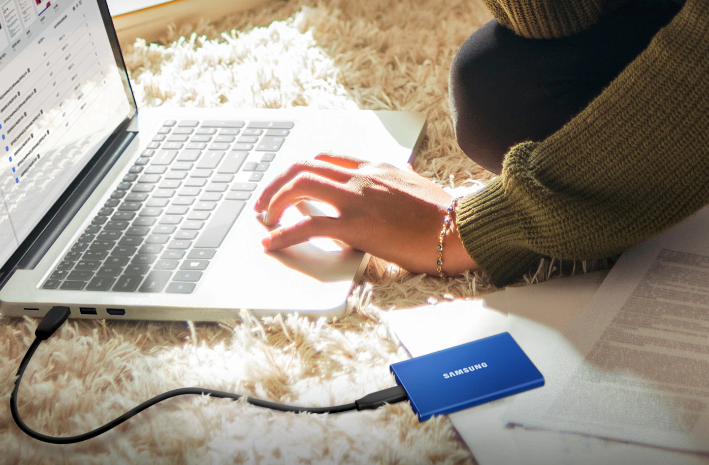 A woman sits on carpeted floor using laptop touchpad with Samsung T7 SSD attached.