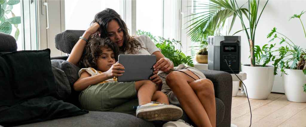 A woman cuddles with her child on the couch, looking at a tablet getting power from an EcoFlow DELTA 2 Portable Power Station.