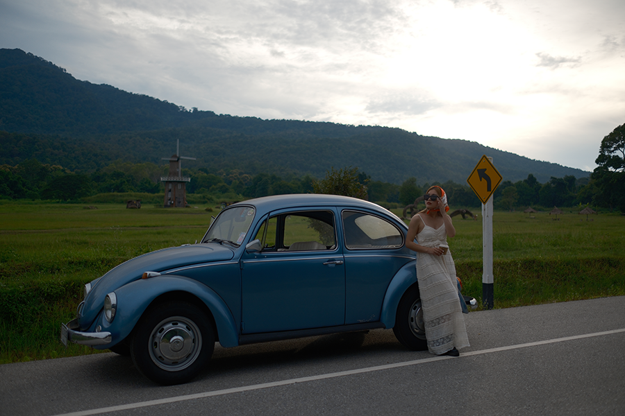 Shot of woman leaning against blue VW bug with small windmill in field behind her, illustrating Nostalgic Neg. film simulation from Fujifilm.