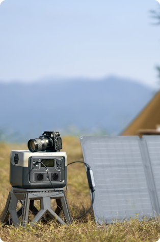 A cropped photo of an EcoFlow RIVER Max 2 portable power station sitting on a stool amongst grass, being charged by a solar panel beside it, with a tent in the background and a camera perched atop the power station.