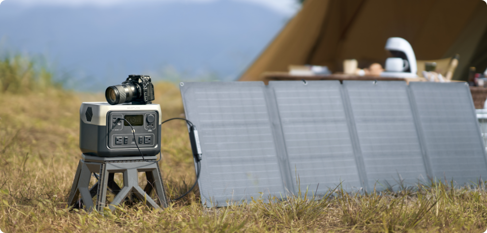 A photo of an EcoFlow RIVER Max 2 portable power station sitting on a stool amongst grass, being charged by a solar panel beside it, with a tent in the background and a camera perched atop the power station.