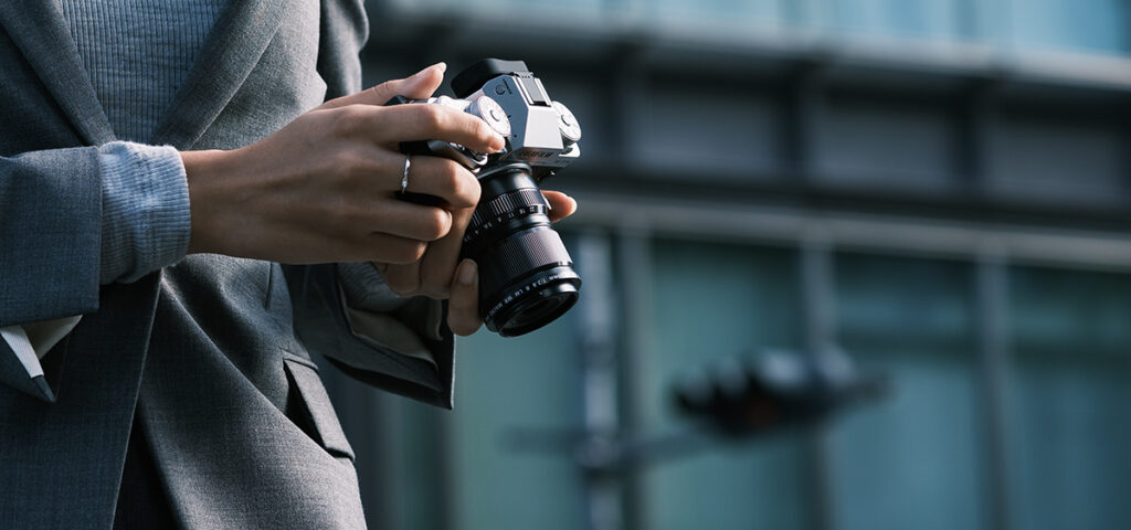 An image showing a woman holding a Fujifilm XF 30mm F2.8 R LM WR Macro lens, tilted up to show the LCD back, with an office building in the background.